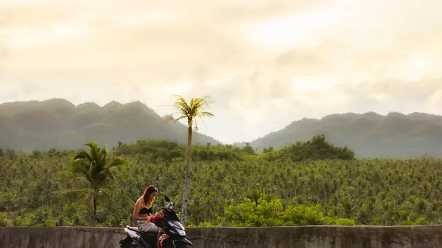 Sweeping vista of a lush coconut palm forest and hills seen from the Coconut Trees View Deck road in Siargao, with a motorbike rider at the roadside.