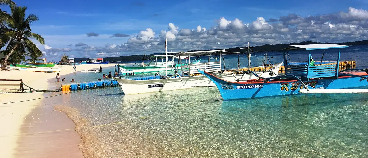 Blue outrigger bangka boats moored in clear shallow water along the white-sand beach of Daku Island, Siargao.