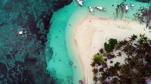 Aerial view of tiny palm-covered Guyam Island with its white sandbar, moored boats and surrounding reef, Siargao.